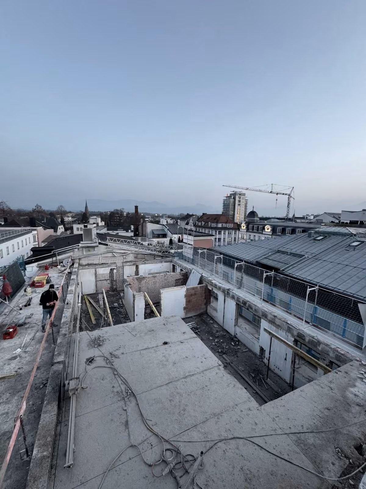Blick über die Dächer bei Dämmerung mit Baukran, Bergen im Hintergrund und einer geöffneten Dachplatte im Vordergrund