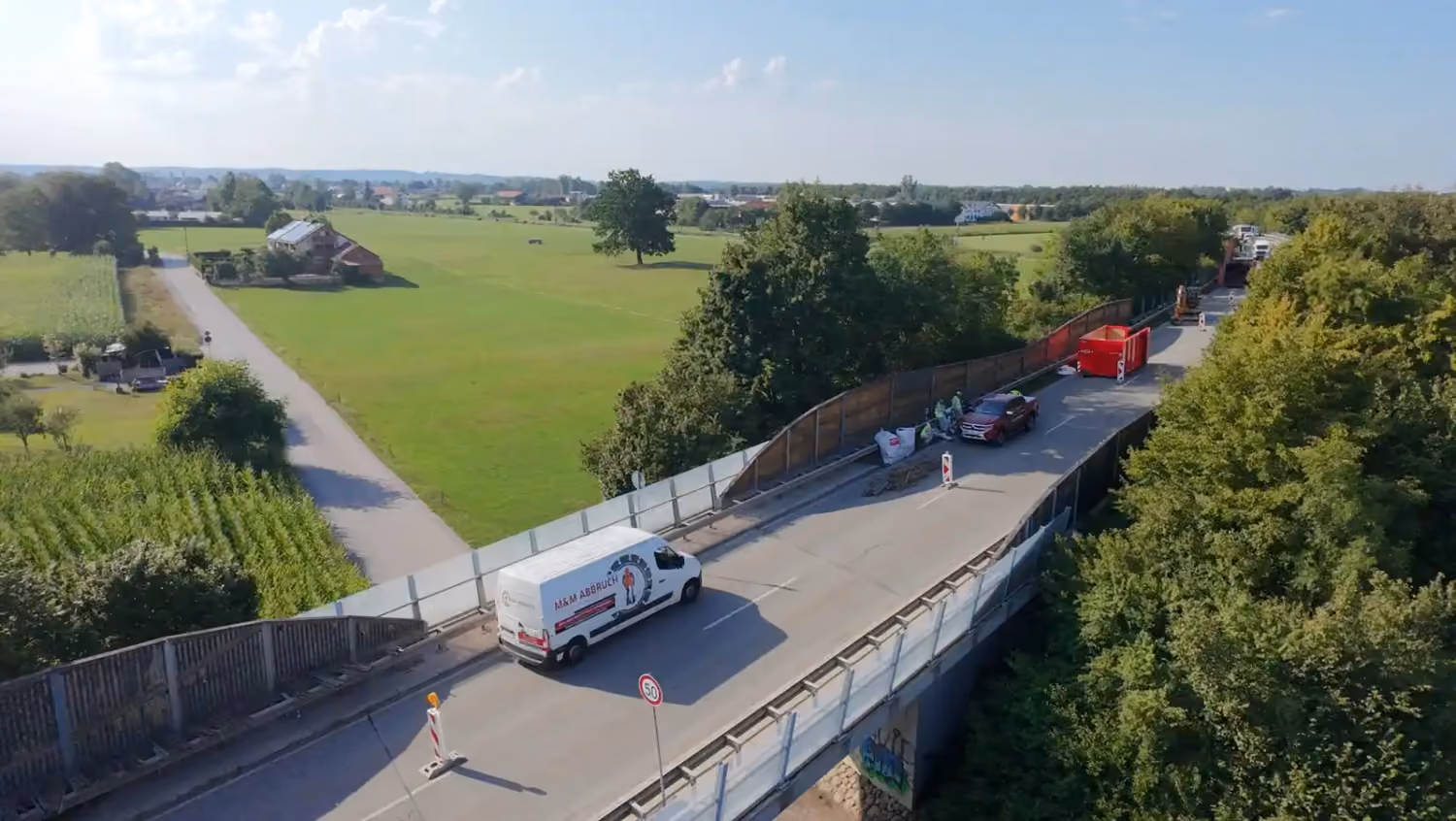 Luftaufnahme einer Baustelle bei Abriss und Erneuerung der Lärmschutzwand einer Straßenbrücke, mit Container und weißem M&M-Abbruch-Transporter
